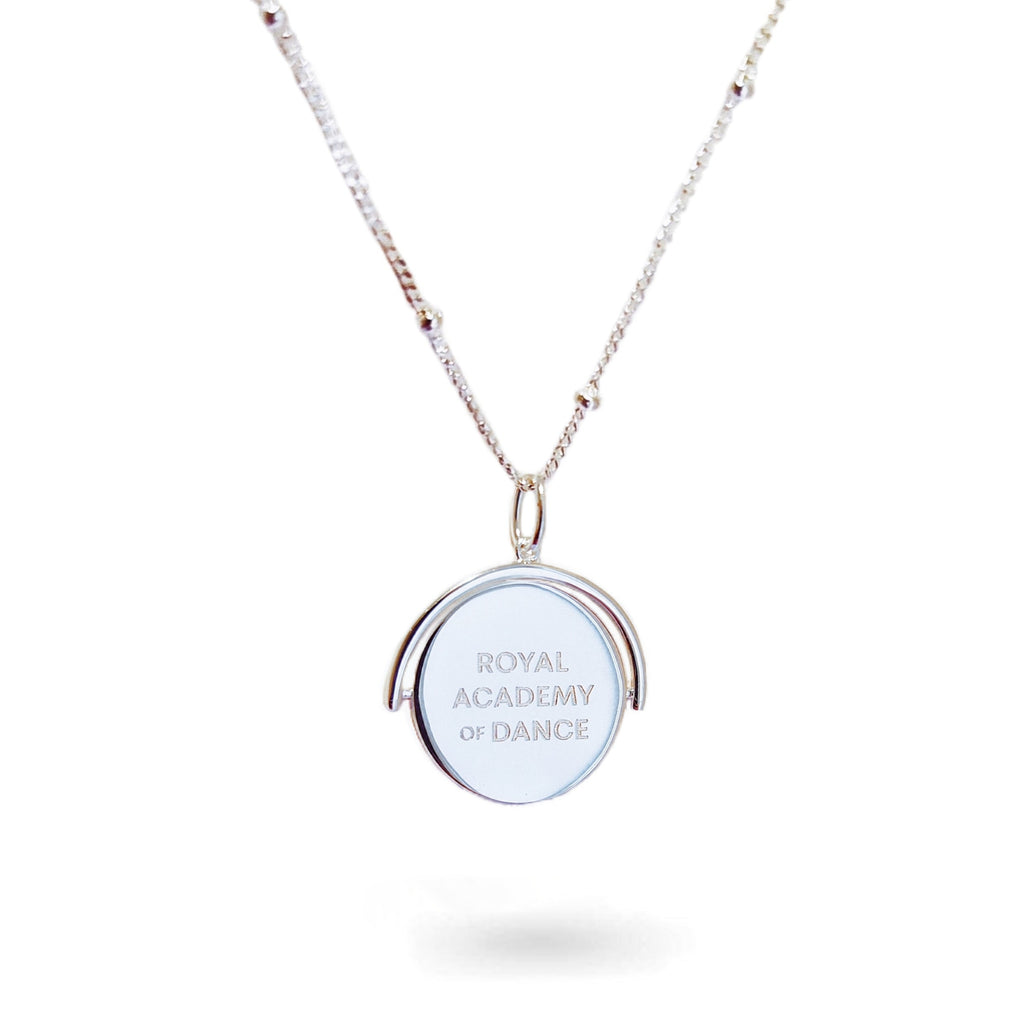 A close-up of a delicate spinning disc pendant on a necklace in stering silver. The circular disc is shown mid-spin, creating a slight blur that hints at its movement. One side features a finely engraved ballerina poised in a graceful arabesque, while the opposite side reveals the formal crest of the Royal Academy of Dance. The disc hangs from a sleek chain, capturing a moment of dynamic elegance where dance and heritage revolve together.
