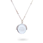 A close-up of a delicate spinning disc pendant on a necklace in stering silver. The circular disc is shown mid-spin, creating a slight blur that hints at its movement. One side features a finely engraved ballerina poised in a graceful arabesque, while the opposite side reveals the formal crest of the Royal Academy of Dance. The disc hangs from a sleek chain, capturing a moment of dynamic elegance where dance and heritage revolve together.