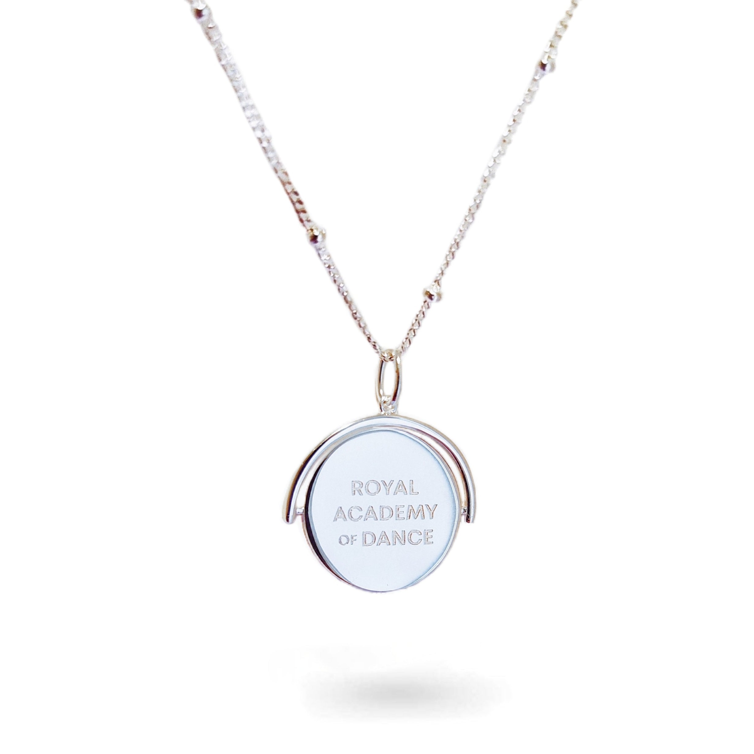 A close-up of a delicate spinning disc pendant on a necklace in stering silver. The circular disc is shown mid-spin, creating a slight blur that hints at its movement. One side features a finely engraved ballerina poised in a graceful arabesque, while the opposite side reveals the formal crest of the Royal Academy of Dance. The disc hangs from a sleek chain, capturing a moment of dynamic elegance where dance and heritage revolve together.