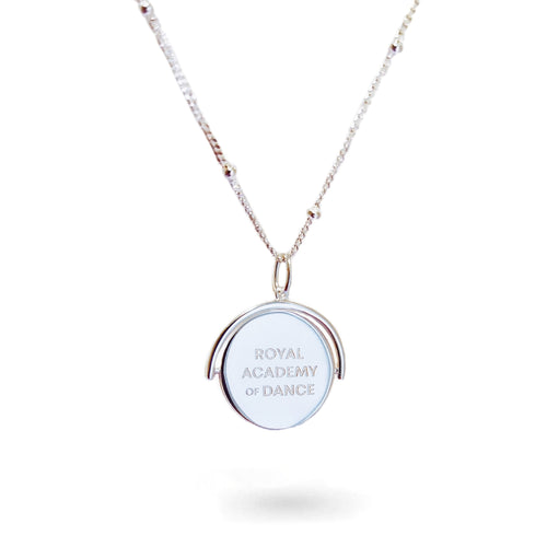 A close-up of a delicate spinning disc pendant on a necklace in stering silver. The circular disc is shown mid-spin, creating a slight blur that hints at its movement. One side features a finely engraved ballerina poised in a graceful arabesque, while the opposite side reveals the formal crest of the Royal Academy of Dance. The disc hangs from a sleek chain, capturing a moment of dynamic elegance where dance and heritage revolve together.
