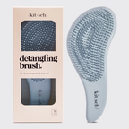A Kit·sch detangling brush shown next to its light pink packaging box on a light gray background. The brush is a pale icy blue with dark blue bristles, and the box features a window displaying the brush inside.