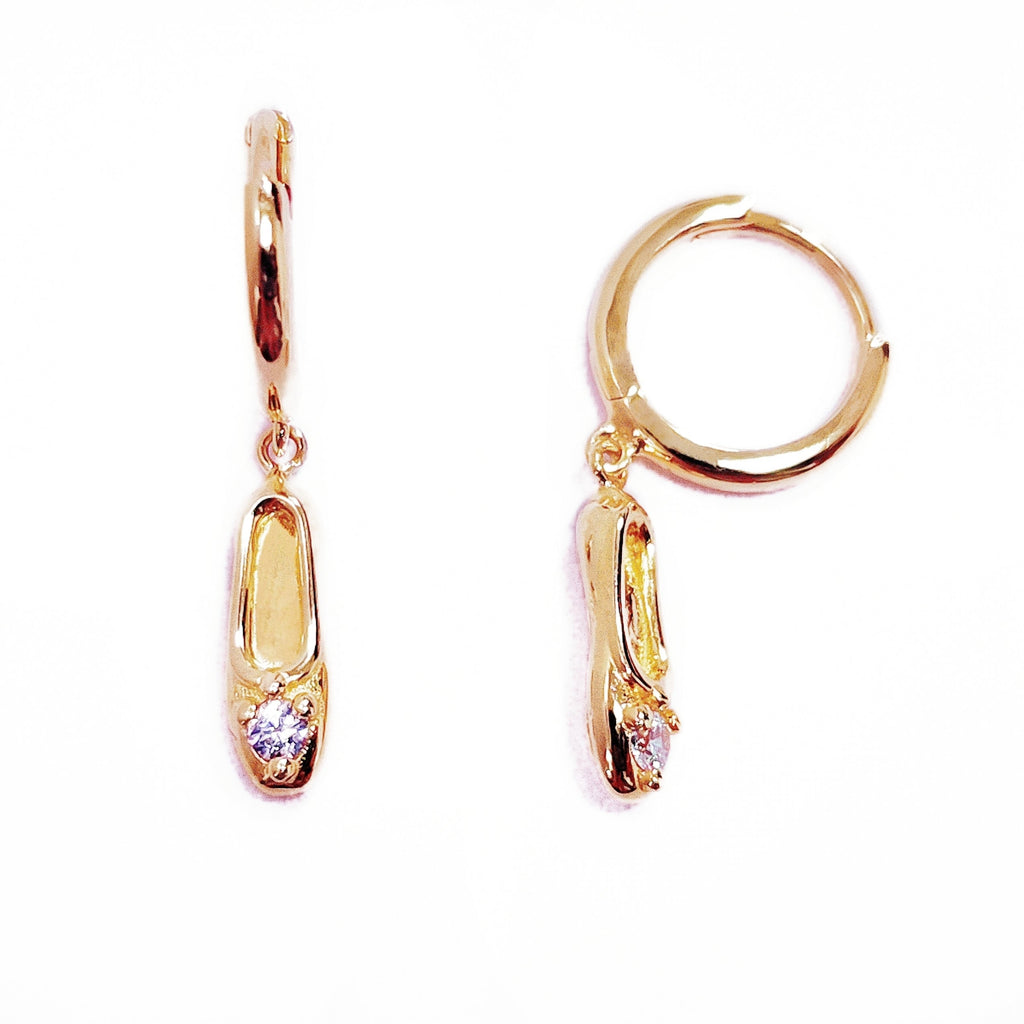 A pair of gold-plated sterling silver huggie hoop earrings, designed to hug the earlobe closely. From the curved base of each hoop dangles a delicate charm in the shape of a classical ballet slipper. The shoe charm is meticulously detailed, featuring sparkling cubic zirconia stone catching the light with each subtle movement. The earrings combine minimalist huggie style with a playful, sparkling tribute to dance. Designed by Royal Academy of Dance. Made in the UK