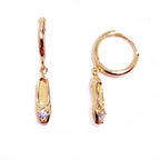 A pair of gold-plated sterling silver huggie hoop earrings, designed to hug the earlobe closely. From the curved base of each hoop dangles a delicate charm in the shape of a classical ballet slipper. The shoe charm is meticulously detailed, featuring sparkling cubic zirconia stone catching the light with each subtle movement. The earrings combine minimalist huggie style with a playful, sparkling tribute to dance. Designed by Royal Academy of Dance. Made in the UK