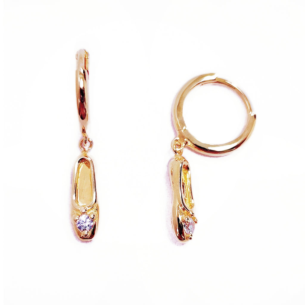A pair of gold-plated sterling silver huggie hoop earrings, designed to hug the earlobe closely. From the curved base of each hoop dangles a delicate charm in the shape of a classical ballet slipper. The shoe charm is meticulously detailed, featuring sparkling cubic zirconia stone catching the light with each subtle movement. The earrings combine minimalist huggie style with a playful, sparkling tribute to dance. Designed by Royal Academy of Dance. Made in the UK