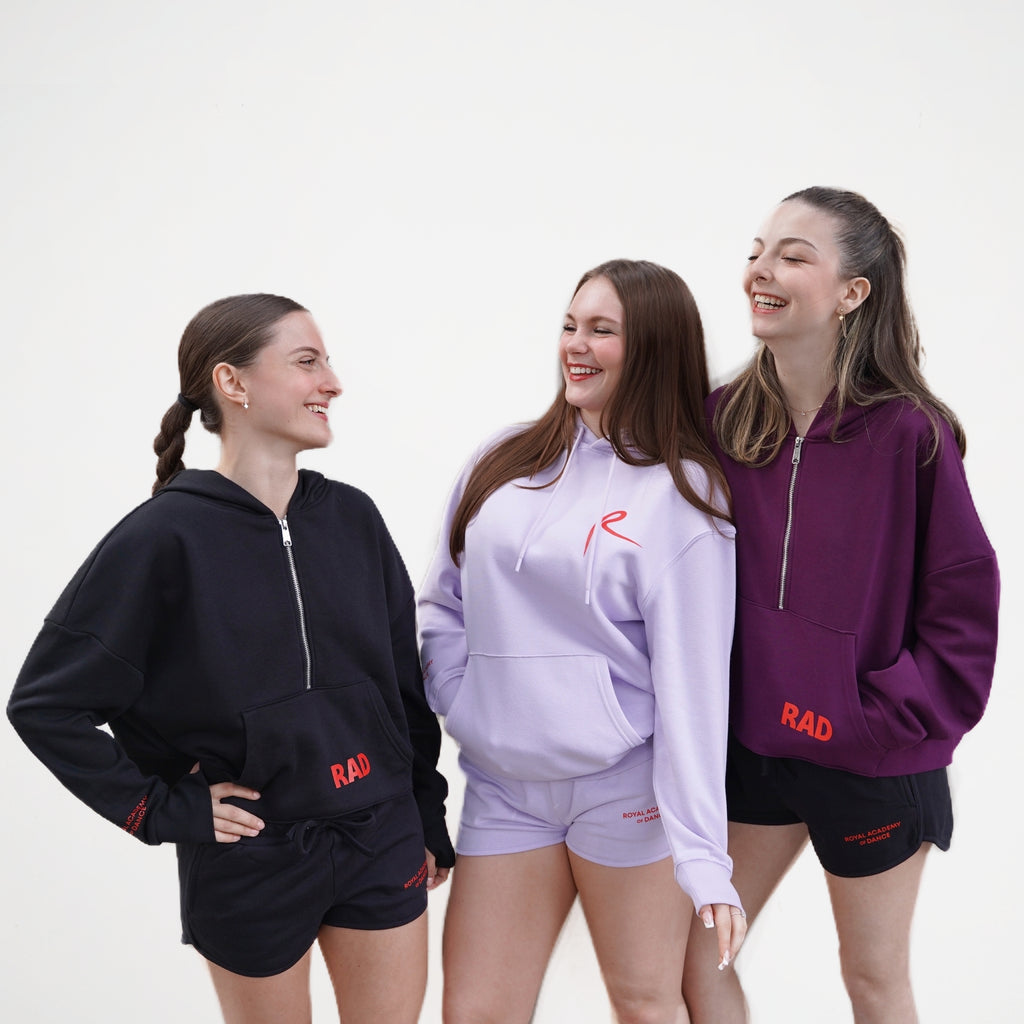  cropped hoodie with a front zip and kangaroo pockets, as well as ribbed cuffs with thumbhole detailing. The hoodie features the text 'RAD' on the front and Royal Academy of Dance on cuff in red. 