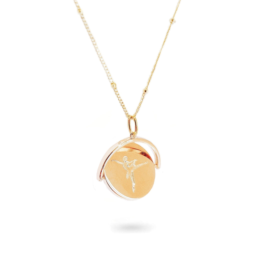 A close-up of a delicate spinning disc pendant on a necklace. The circular disc is shown mid-spin, creating a slight blur that hints at its movement. One side features a finely engraved ballerina poised in a graceful arabesque, while the opposite side reveals the formal crest of the Royal Academy of Dance. The disc hangs from a sleek chain, capturing a moment of dynamic elegance where dance and heritage revolve together.