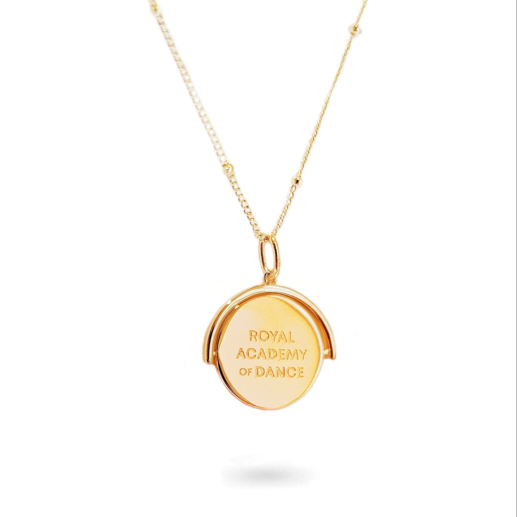 A close-up of a delicate spinning disc pendant on a necklace in gold plated silver. The circular disc is shown mid-spin, creating a slight blur that hints at its movement. One side features a finely engraved ballerina poised in a graceful arabesque, while the opposite side reveals the formal crest of the Royal Academy of Dance. The disc hangs from a sleek chain, capturing a moment of dynamic elegance where dance and heritage revolve together.