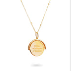 A close-up of a delicate spinning disc pendant on a necklace in gold plated silver. The circular disc is shown mid-spin, creating a slight blur that hints at its movement. One side features a finely engraved ballerina poised in a graceful arabesque, while the opposite side reveals the formal crest of the Royal Academy of Dance. The disc hangs from a sleek chain, capturing a moment of dynamic elegance where dance and heritage revolve together.