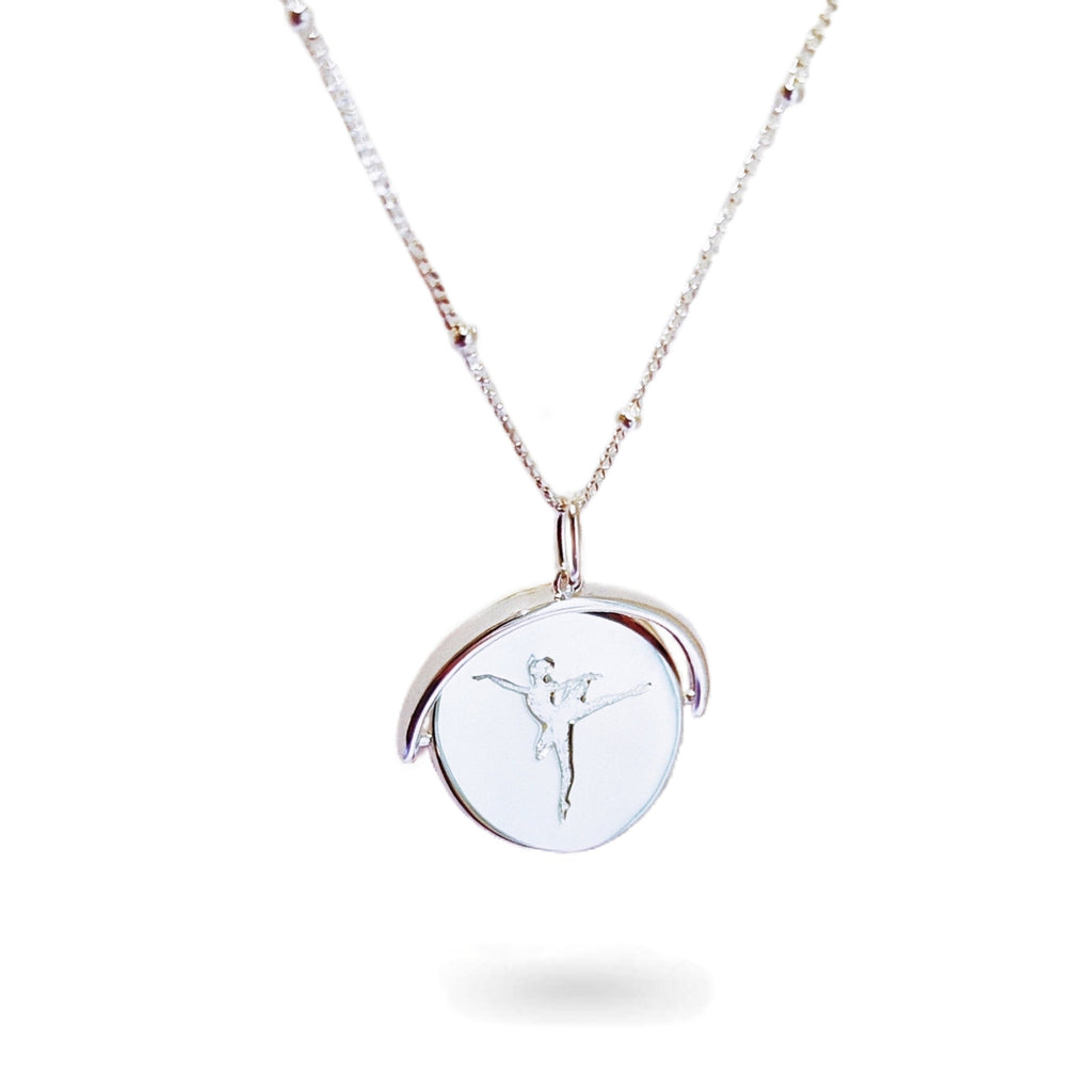 A close-up of a delicate spinning disc pendant on a necklace. The circular disc is shown mid-spin, creating a slight blur that hints at its movement. One side features a finely engraved ballerina poised in a graceful arabesque, while the opposite side reveals the formal crest of the Royal Academy of Dance. The disc hangs from a sleek chain, capturing a moment of dynamic elegance where dance and heritage revolve together.