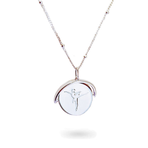 A close-up of a delicate spinning disc pendant on a necklace. The circular disc is shown mid-spin, creating a slight blur that hints at its movement. One side features a finely engraved ballerina poised in a graceful arabesque, while the opposite side reveals the formal crest of the Royal Academy of Dance. The disc hangs from a sleek chain, capturing a moment of dynamic elegance where dance and heritage revolve together.