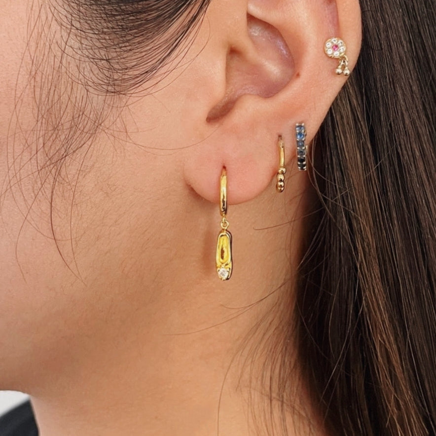 A pair of gold-plated sterling silver huggie hoop earrings, designed to hug the earlobe closely. From the curved base of each hoop dangles a delicate charm in the shape of a classical ballet slipper. The shoe charm is meticulously detailed, featuring sparkling cubic zirconia stone catching the light with each subtle movement. The earrings combine minimalist huggie style with a playful, sparkling tribute to dance. Designed by Royal Academy of Dance. Made in the UK