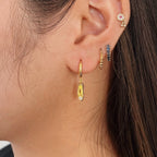A pair of gold-plated sterling silver huggie hoop earrings, designed to hug the earlobe closely. From the curved base of each hoop dangles a delicate charm in the shape of a classical ballet slipper. The shoe charm is meticulously detailed, featuring sparkling cubic zirconia stone catching the light with each subtle movement. The earrings combine minimalist huggie style with a playful, sparkling tribute to dance. Designed by Royal Academy of Dance. Made in the UK