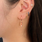 A pair of rose-gold-plated sterling silver huggie hoop earrings, designed to hug the earlobe closely. From the curved base of each hoop dangles a delicate charm in the shape of a classical ballet slipper. The shoe charm is meticulously detailed, featuring sparkling cubic zirconia stone, catching the light with each subtle movement. The earrings combine minimalist huggie style with a playful, sparkling tribute to dance. Designed by Royal Academy of Dance. Made in the UK