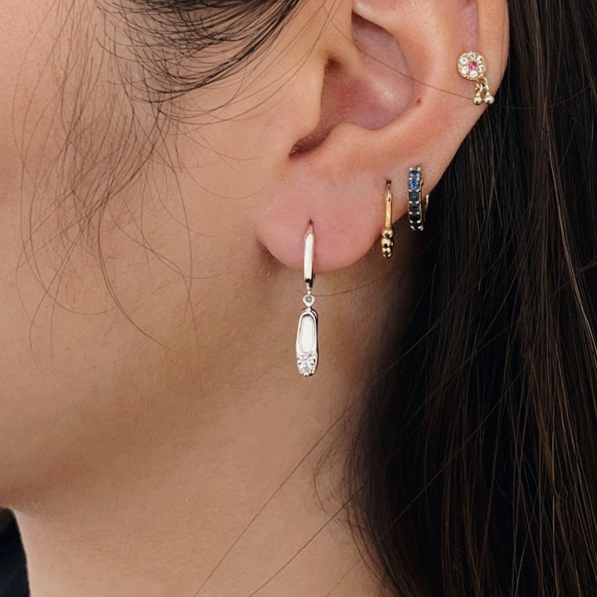 A pair of sterling silver huggie hoop earrings, designed to hug the earlobe closely. From the curved base of each hoop dangles a delicate charm in the shape of a classical ballet slipper. The shoe charm is meticulously detailed, featuring sparkling cubic zirconia stone, catching the light with each subtle movement. The earrings combine minimalist huggie style with a playful, sparkling tribute to dance. Designed by Royal Academy of Dance. Made in the UK