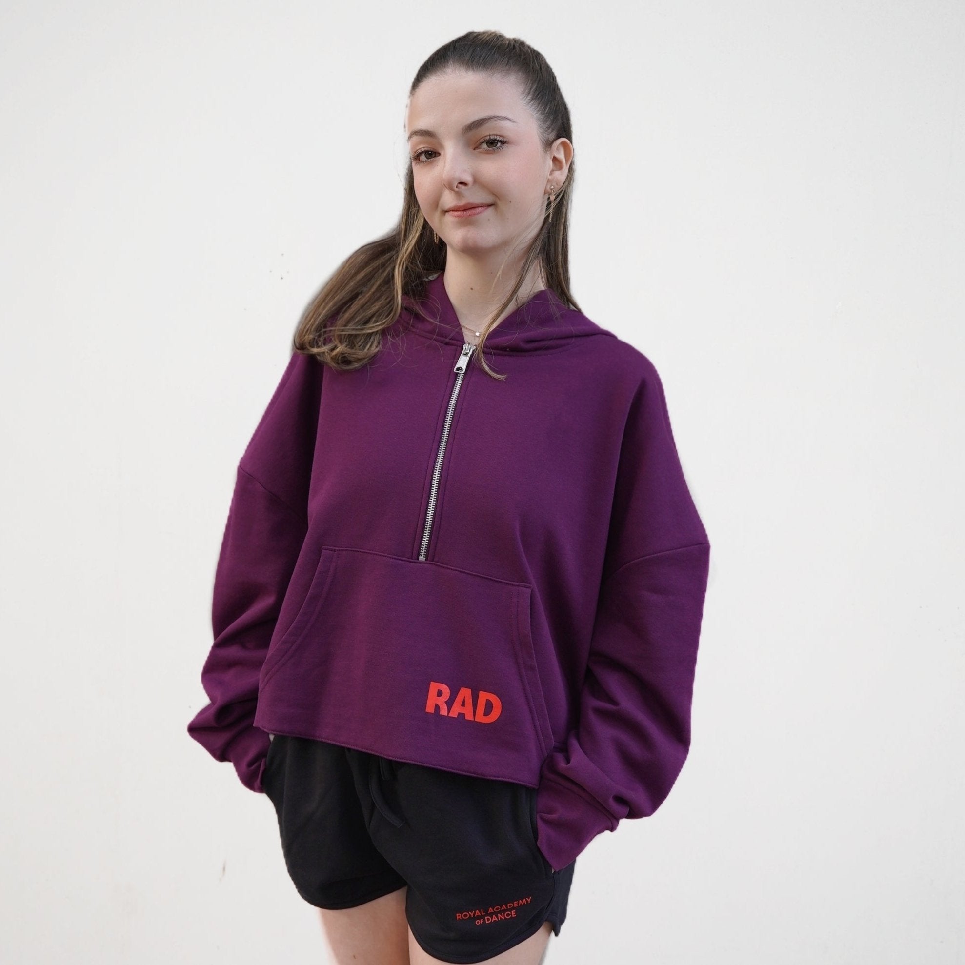Mulberry  cropped hoodie with a front zip and kangaroo pockets, as well as ribbed cuffs with thumbhole detailing. The hoodie features the text 'RAD' on the front and Royal Academy of Dance on cuff in red. 
