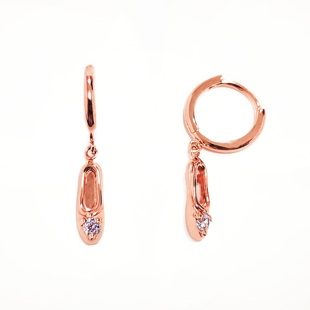 A pair of rose-gold-plated sterling silver huggie hoop earrings, designed to hug the earlobe closely. From the curved base of each hoop dangles a delicate charm in the shape of a classical ballet slipper. The shoe charm is meticulously detailed, featuring sparkling cubic zirconia stones that adorn the ribbon and sole, catching the light with each subtle movement. The earrings combine minimalist huggie style with a playful, sparkling tribute to dance. Designed by Royal Academy of Dance. Made in the UK