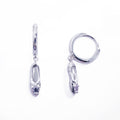 A pair of sterling silver huggie hoop earrings, designed to hug the earlobe closely. From the curved base of each hoop dangles a delicate charm in the shape of a classical ballet slipper. The shoe charm is meticulously detailed, featuring sparkling cubic zirconia stone, catching the light with each subtle movement. The earrings combine minimalist huggie style with a playful, sparkling tribute to dance. Designed by Royal Academy of Dance. Made in the UK