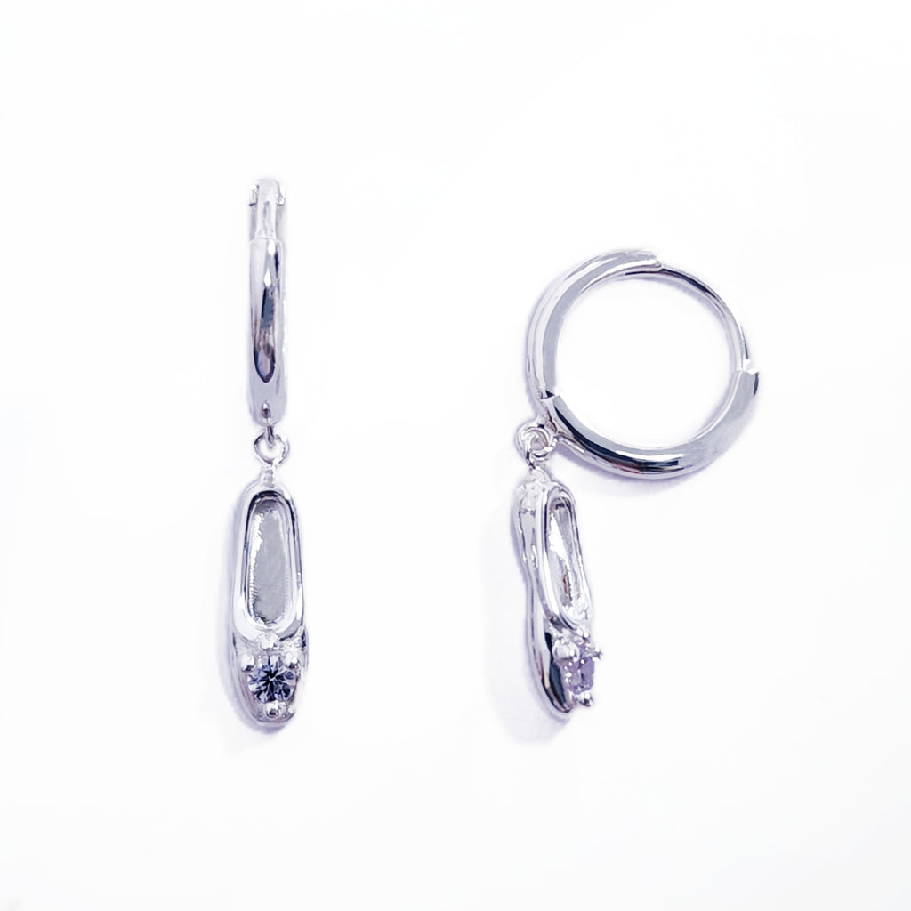 A pair of sterling silver huggie hoop earrings, designed to hug the earlobe closely. From the curved base of each hoop dangles a delicate charm in the shape of a classical ballet slipper. The shoe charm is meticulously detailed, featuring sparkling cubic zirconia stone, catching the light with each subtle movement. The earrings combine minimalist huggie style with a playful, sparkling tribute to dance. Designed by Royal Academy of Dance. Made in the UK