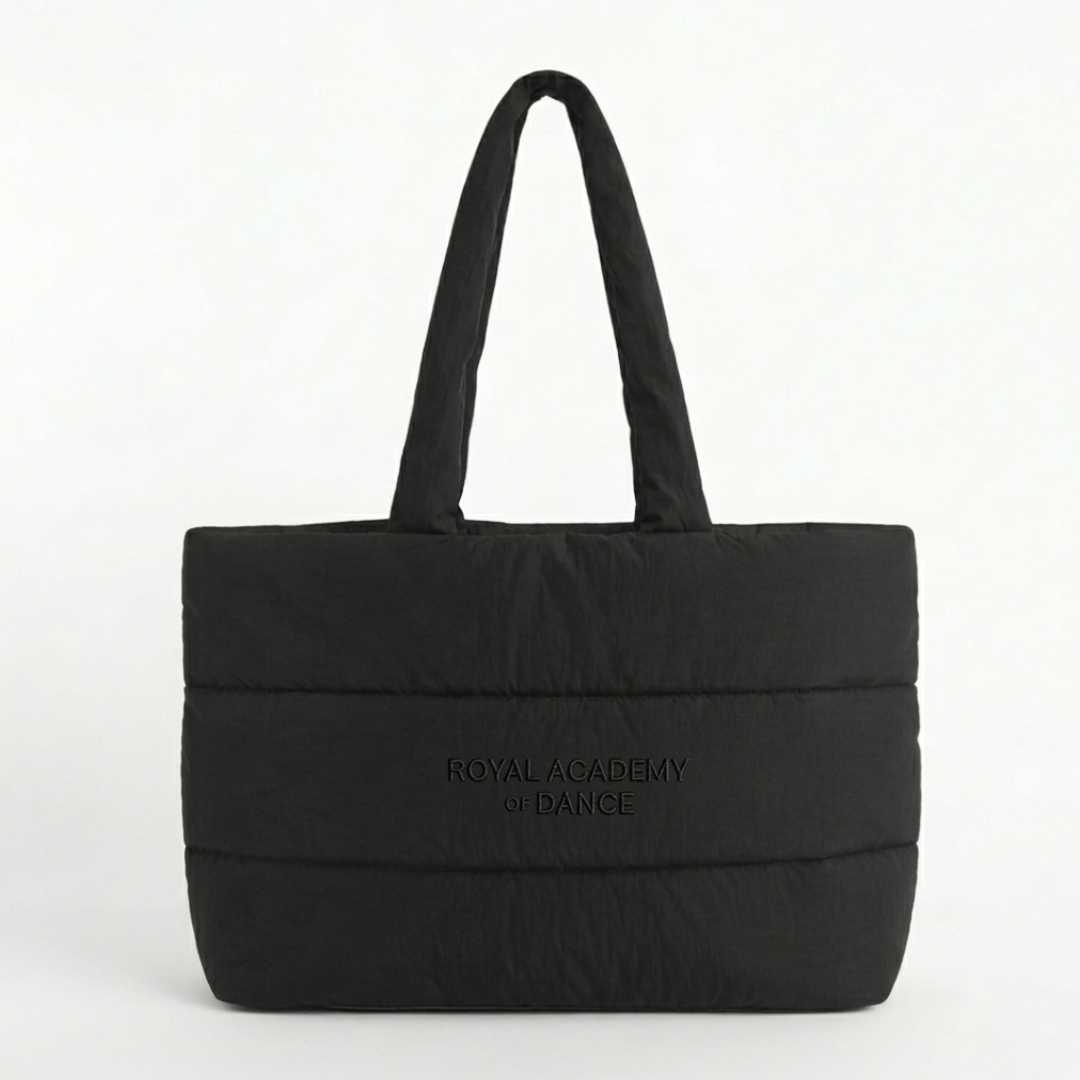 This oversized black quilted puffer tote bag features the Royal Academy of Dance logo, blending a professional ballet aesthetic with new streetwear trends. Designed as a spacious dance bag for dancers on the go, the lightweight padded nylon and soft cloud-like texture provide a stylish studio-to-street look. The large interior easily holds pointe shoes and warm-ups, making it a functional yet trendy athleisure accessory for rehearsals, travel, or everyday use