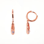 A pair of rose-gold-plated sterling silver huggie hoop earrings, designed to hug the earlobe closely. From the curved base of each hoop dangles a delicate charm in the shape of a classical ballet slipper. The shoe charm is meticulously detailed, featuring sparkling cubic zirconia stone, catching the light with each subtle movement. The earrings combine minimalist huggie style with a playful, sparkling tribute to dance. Designed by Royal Academy of Dance. Made in the UK