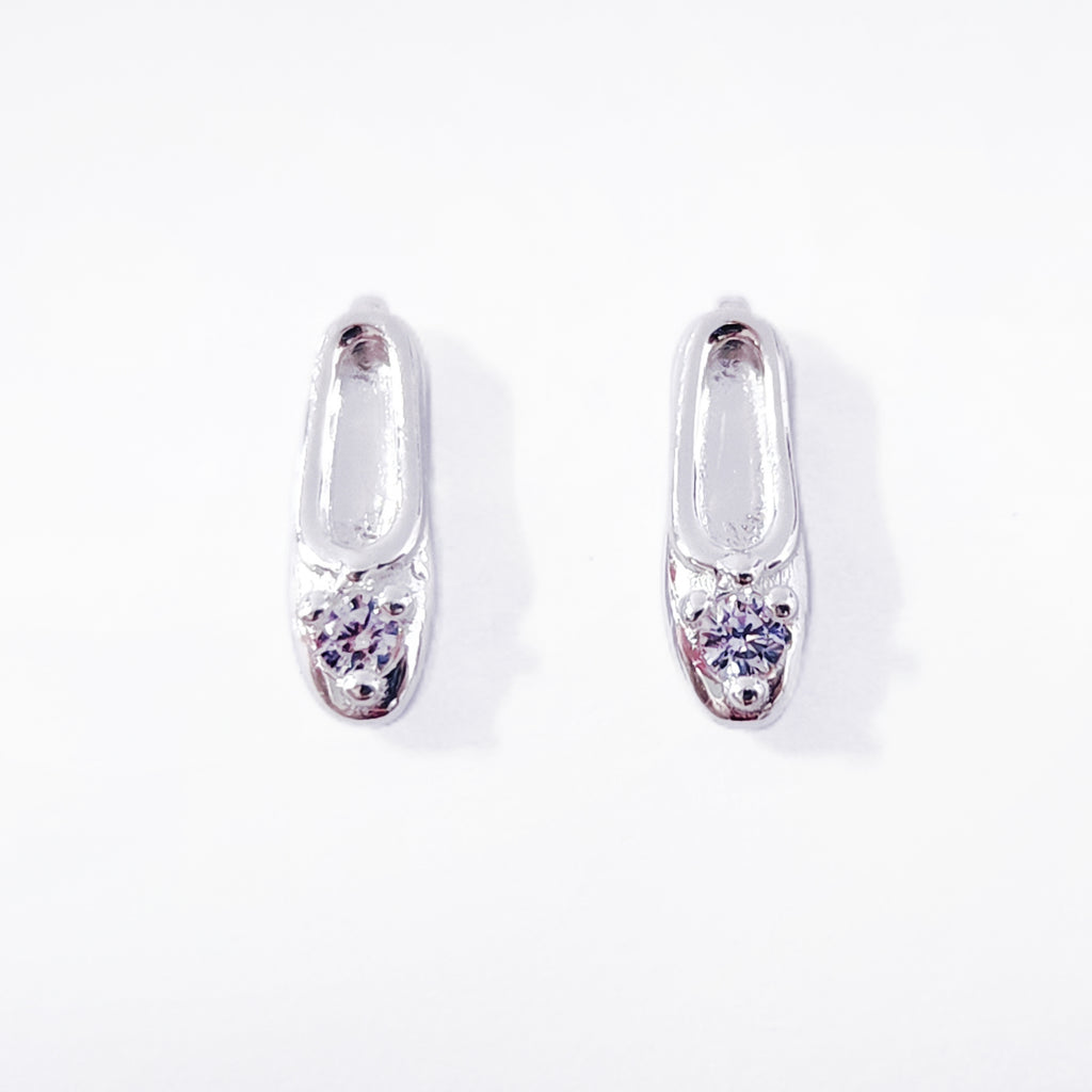 A pair of classic sterling silver stud earrings, each crafted in the shape of a petite, elegant ballet slipper. A single, brilliant cubic zirconia is set at top of the shoe, acting as a sparkling focal point. These dainty studs offer a subtle and refined nod to ballet, perfect for everyday wear. Designed by Royal Academy of Dance. Made in the UK.