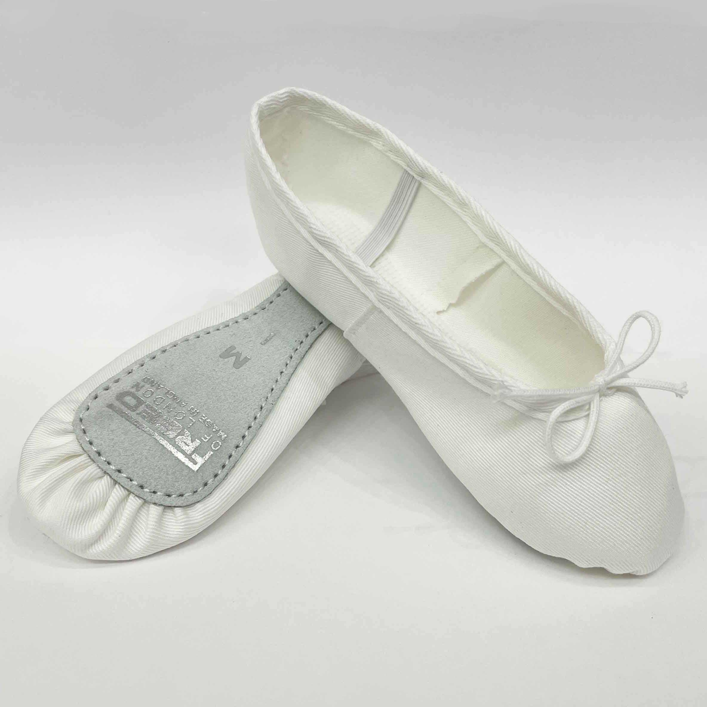Freed of London split sole white canvas ballet shoes, part of the official Royal Academy of Dance exam uniform. The durable matte canvas upper is breathable and flexible. One shoe is propped up, revealing the hand-stitched grey suede split sole patch for enhanced flexible support and arch articulation. The professional-grade ballet flat features pre-sewn elastic straps and a neat front drawstring bow, designed for a sleek, breathable fit for dancers of all grades.